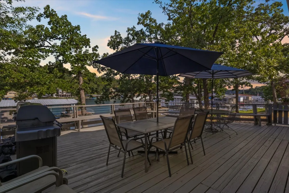 A spacious outdoor setup featuring a dining table with chairs, large umbrellas for shade, a grill for barbecues, and a scenic view of the water beyond. The surrounding trees add privacy and natural beauty, while the arrangement invites group meals and social events under the open sky.