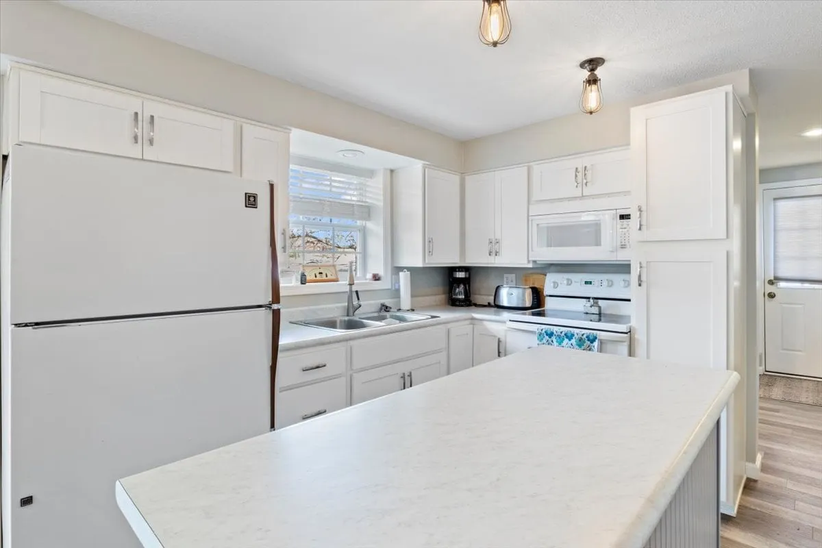 A bright, functional kitchen featuring a spacious island, white cabinetry, and modern appliances. Natural light streams in through the window, creating a cheerful and inviting space ideal for cooking and gathering. The setup highlights ample counter space and a welcoming atmosphere for shared culinary experiences.