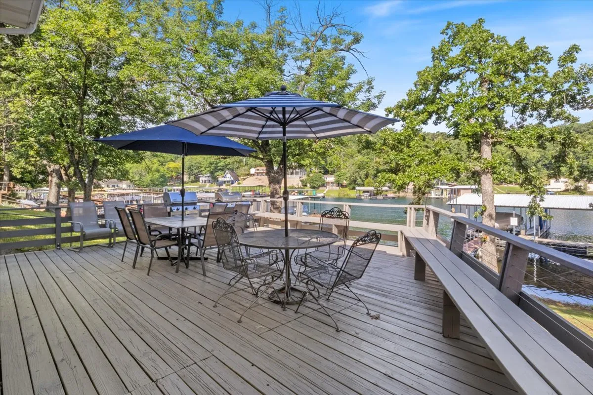 A vibrant outdoor dining table sits beneath a large umbrella, overlooking the tranquil water. The scene is framed by lush trees and captures the ambiance of lakeside al fresco dining. The table is set for gatherings, with the sparkling water just steps away, suggesting a lively yet relaxed environment ideal for socializing.