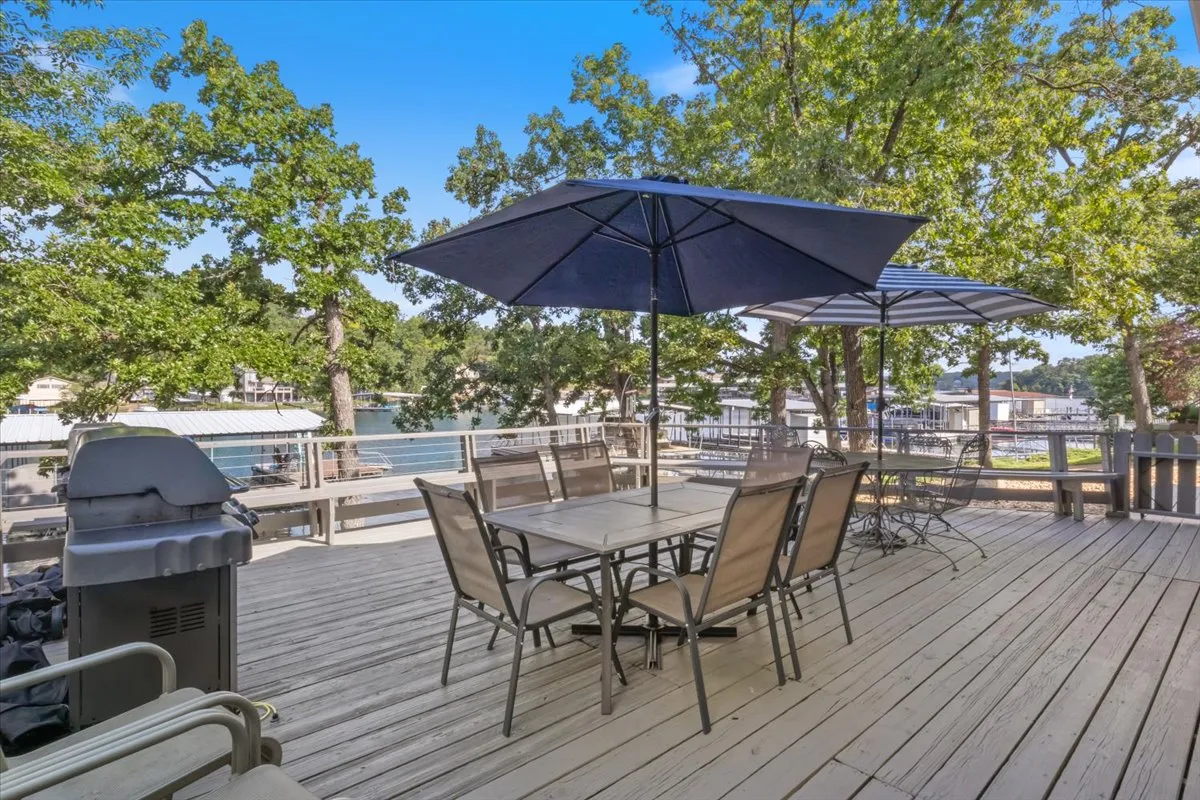 An outdoor deck scene featuring a dining table with chairs, large umbrellas for shade, a visible grill, and a scenic view of the lake framed by trees. The setup is ready for outdoor meals, barbecues, and enjoying the lakeside ambiance, blending al fresco dining with beautiful natural surroundings.