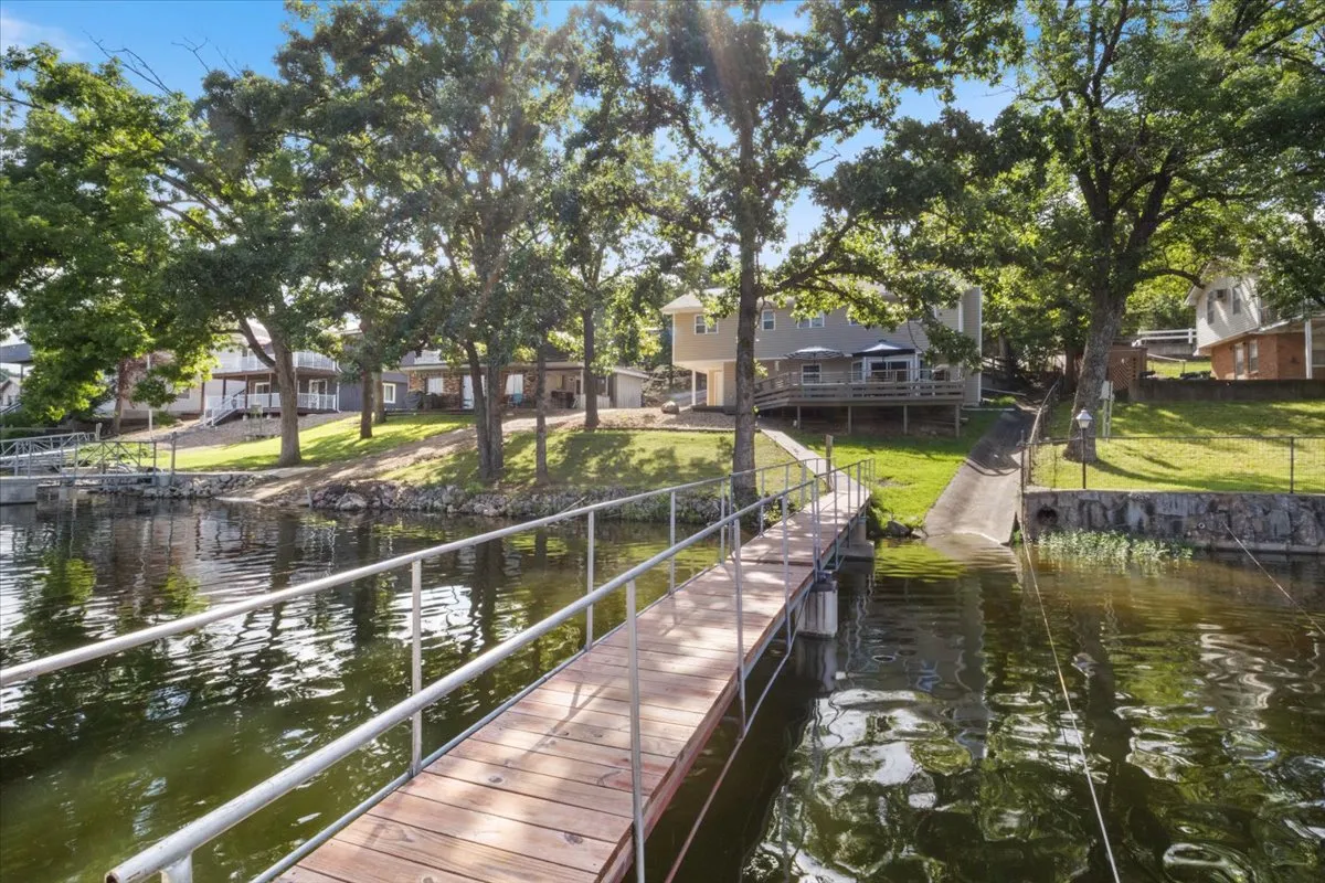 A tranquil lakeside scene featuring a private wooden dock stretching into calm water, surrounded by lush green trees and a manicured lawn. The image shows enough space for swimming, fishing, and launching a boat, with nearby houses visible, suggesting a quiet cove setting ideal for families.