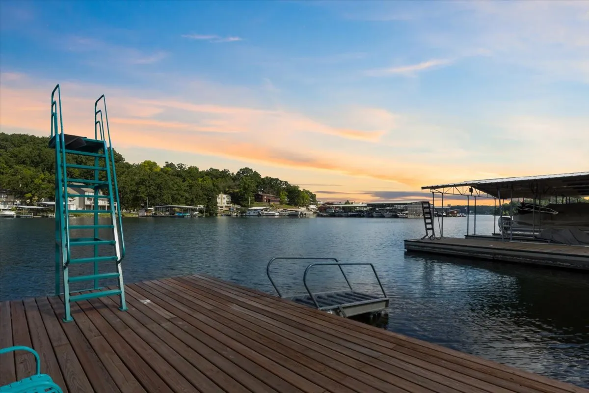 The image showcases a serene lakeside setting at sunset, featuring a wooden dock extending into calm water. A sturdy dive ladder is visible at the end of the dock, inviting swimming and relaxation. The sky glows with warm sunset colors, while the peaceful cove hints at privacy and tranquility. The scene suggests easy access for boating, fishing, and water fun, with the expansive lake stretching out beyond the dock.